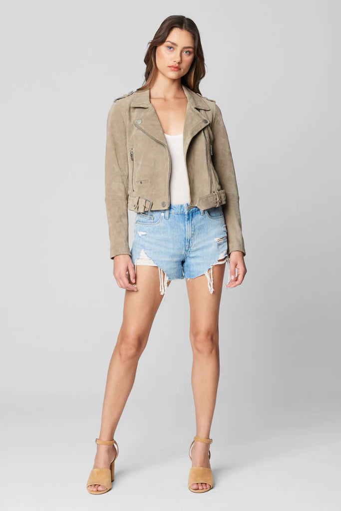 Woman wearing a beige jacket, white top, and denim shorts on a gray background