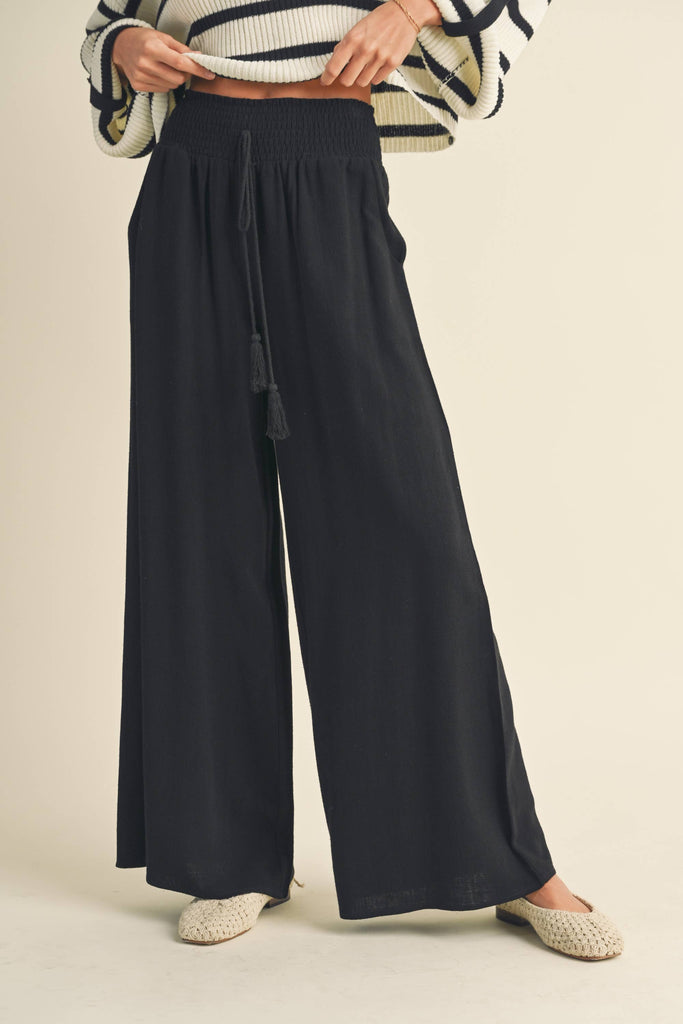 Woman wearing black wide-leg pants with smocked waistband and tassel drawstring.