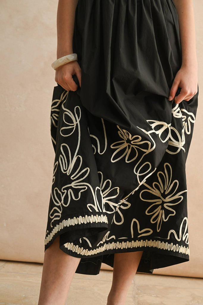 Woman wearing a black spaghetti-strap maxi dress with ivory floral embroidery and scalloped trim detail.