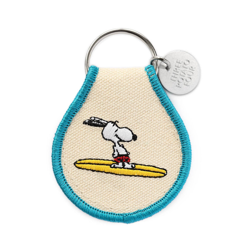 Peanuts® - Snoopy Surf Keychain with an embroidered design of Snoopy leaning on a yellow and red surfboard, framed with blue trim, and a metal “Three Potato Four” tag.