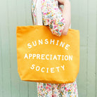 Yellow canvas tote bag with “Sunshine Appreciation Society” printed in white, held up by a hand in front of a sage green wall