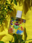 Maison Matine Nature Insolente perfume bottle with colorful leaf and beetle illustrations, placed on a vibrant yellow fabric and surrounded by green foliage.