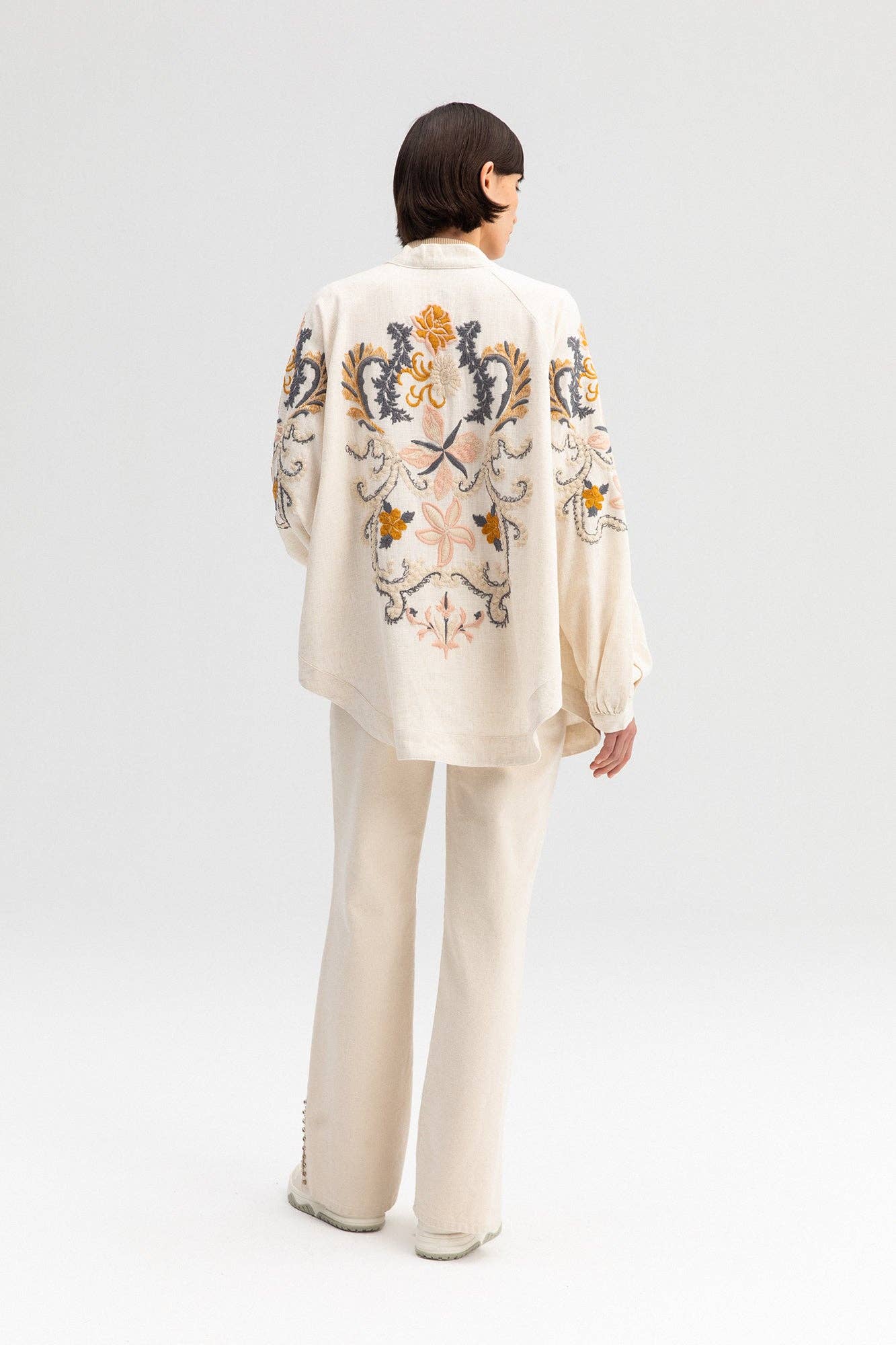 Back view of a cream-colored kimono jacket with colorful floral embroidery, worn with matching light trousers.