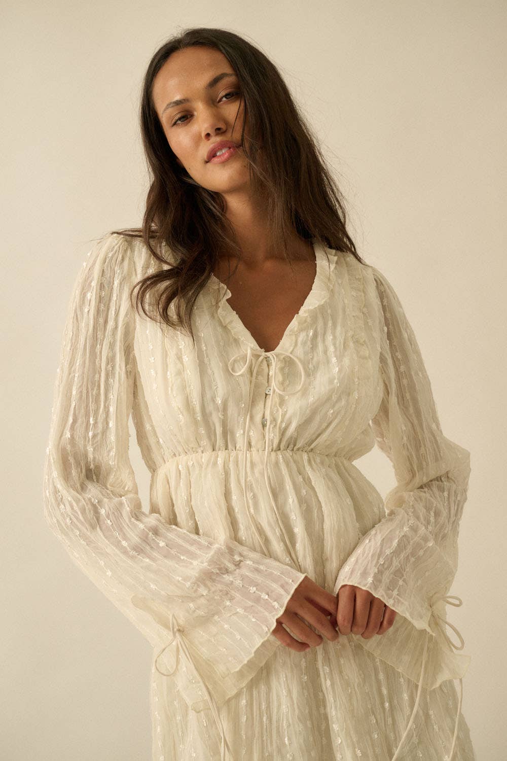 Woman wearing a cream mini dress with sheer long sleeves, button front, and tie details.
