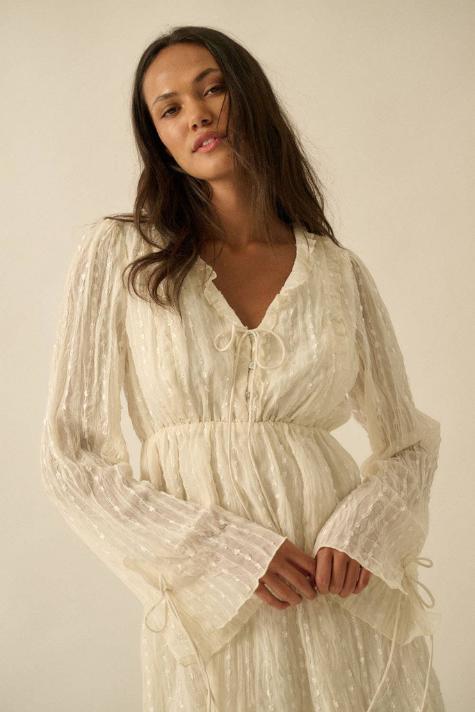 Woman wearing a cream mini dress with sheer long sleeves, button front, and tie details.