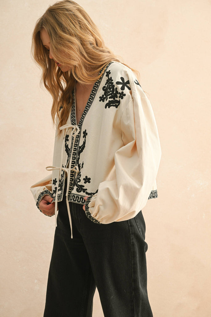 Woman wearing cream blouse with black floral embroidery, lace-up front, and long billowy sleeves.