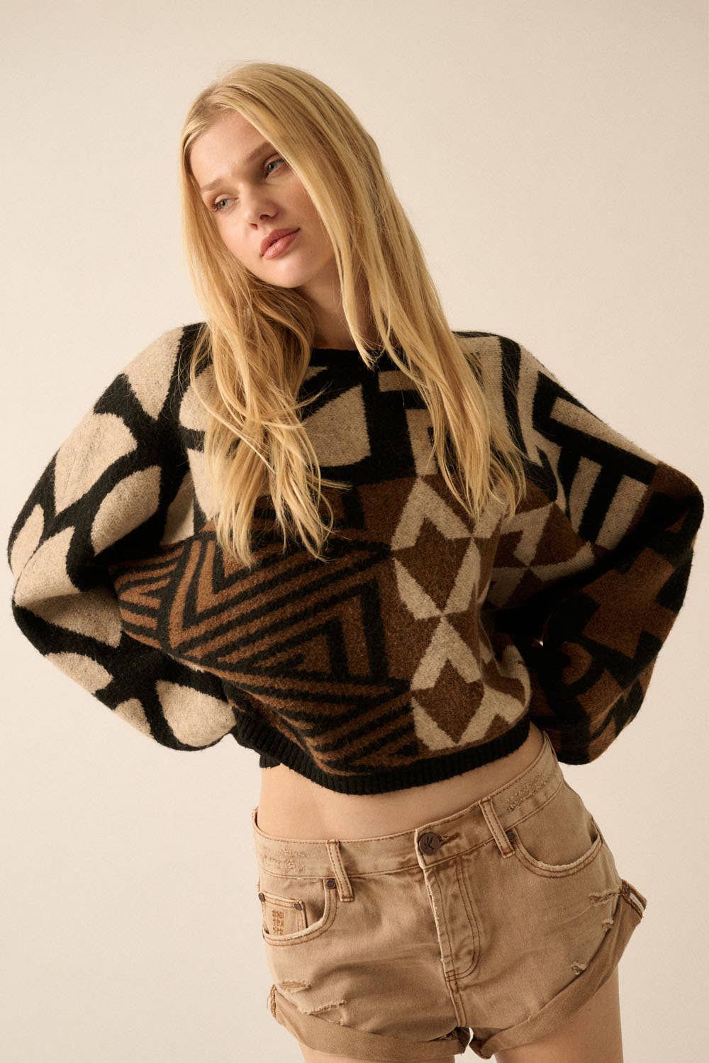 Model wearing a patterned knit sweater with bold geometric designs in neutral and contrasting tones, styled with casual bottoms.