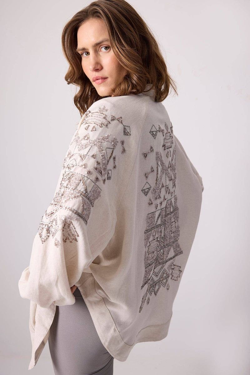 Woman wearing a cream linen kimono jacket with silver sequin embroidery on the sleeves and back.