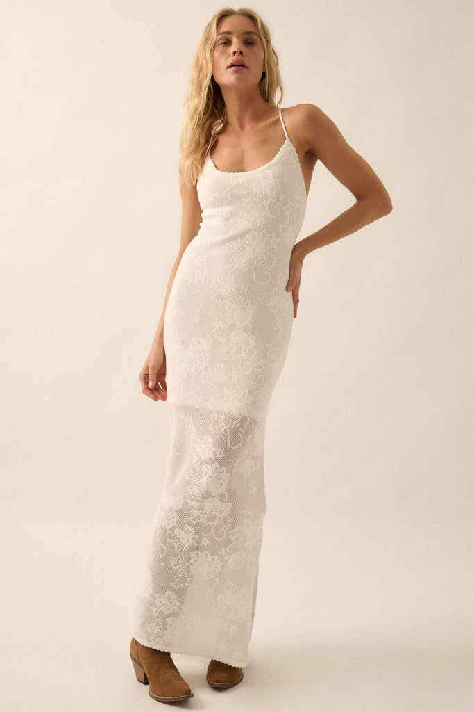 Woman wearing a sheer white lace maxi dress with spaghetti straps and floral detailing.