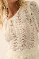 Woman wearing a beige knit romper with bell sleeves, scalloped hems, and open-weave detailing.