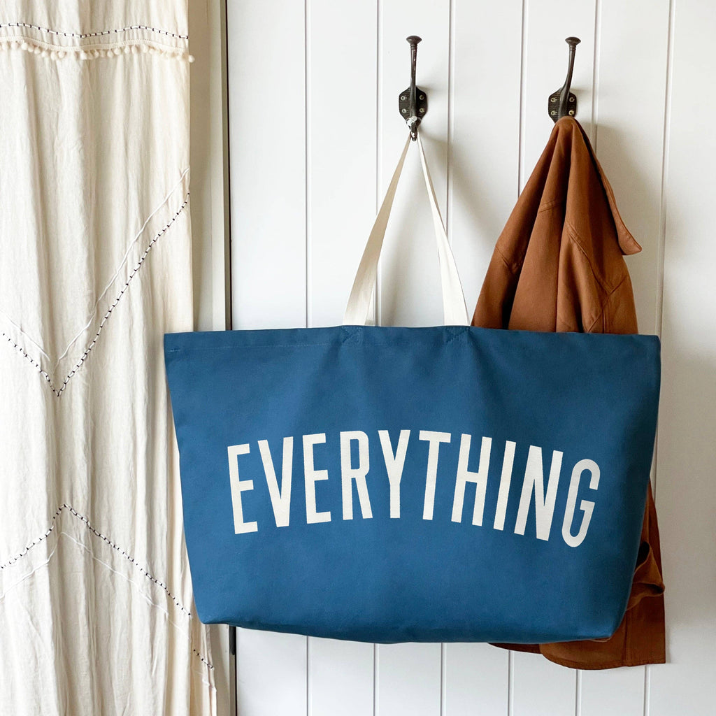 Oversized navy blue tote bag with cream handles and bold white “EVERYTHING” text, held by a hand against a light wood background