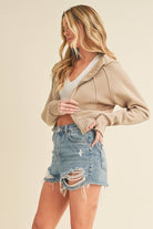 Model wearing a cropped zip-up hoodie with adjustable hood and ribbed cuffs, paired with distressed denim shorts.