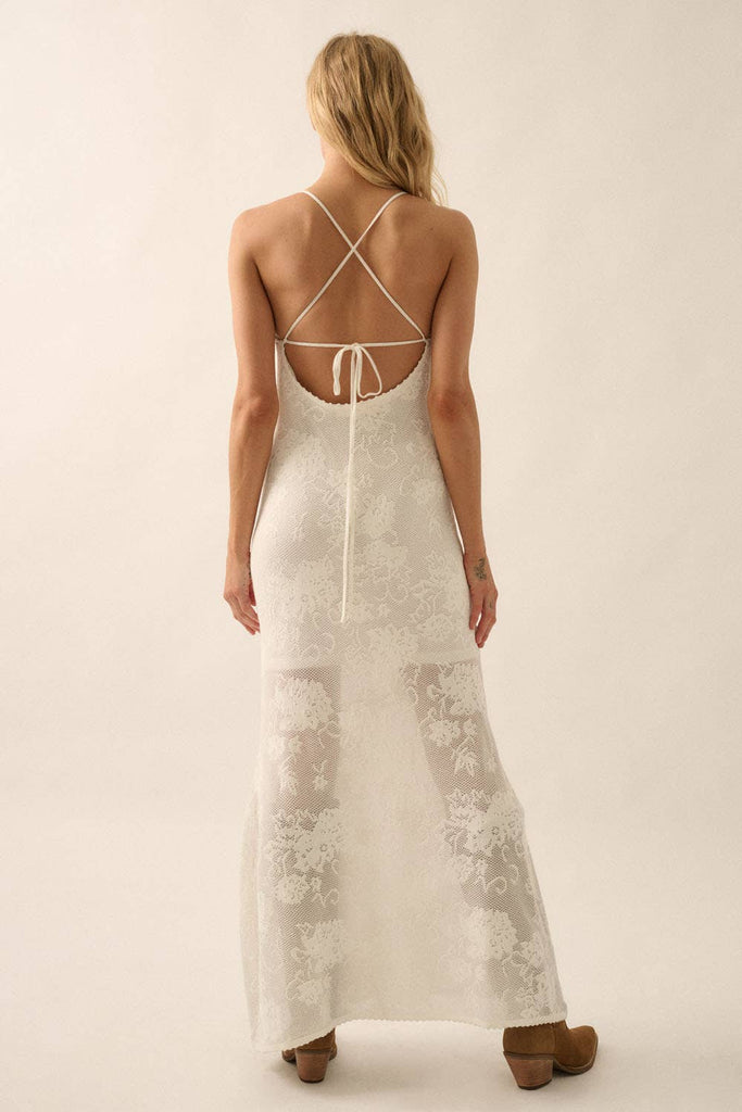 Woman wearing a sheer white lace maxi dress with spaghetti straps and floral detailing.