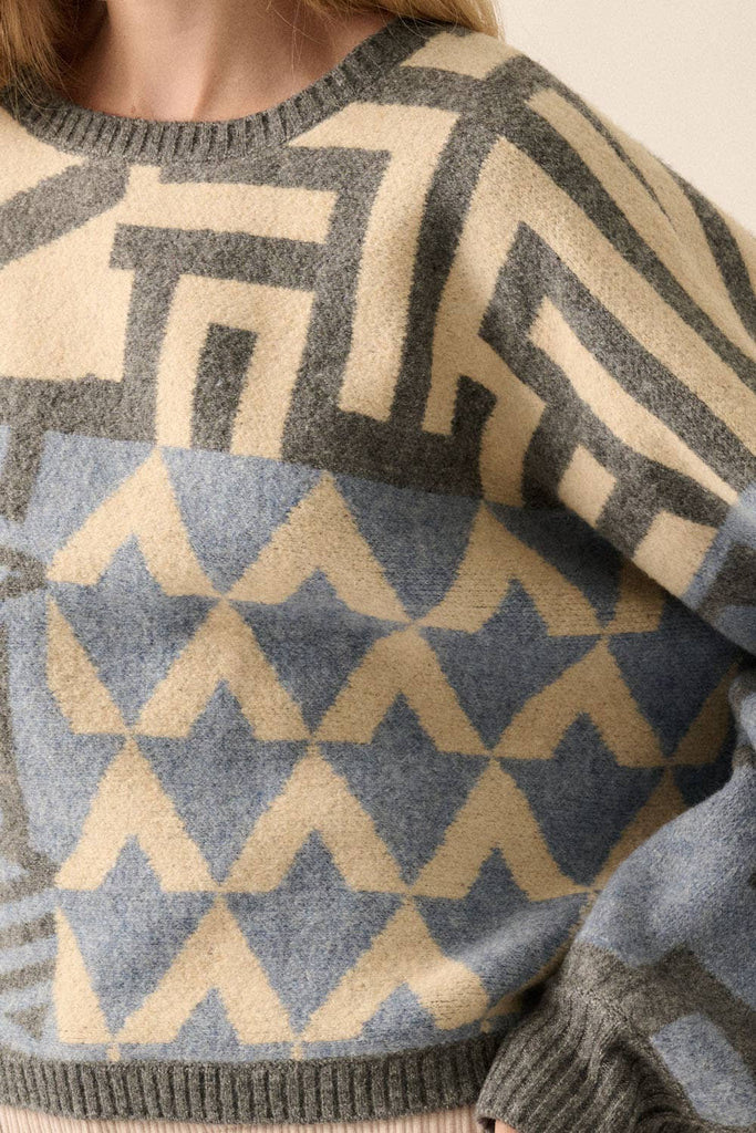 Model wearing a patterned knit sweater with bold geometric designs in neutral and contrasting tones, styled with casual bottoms.