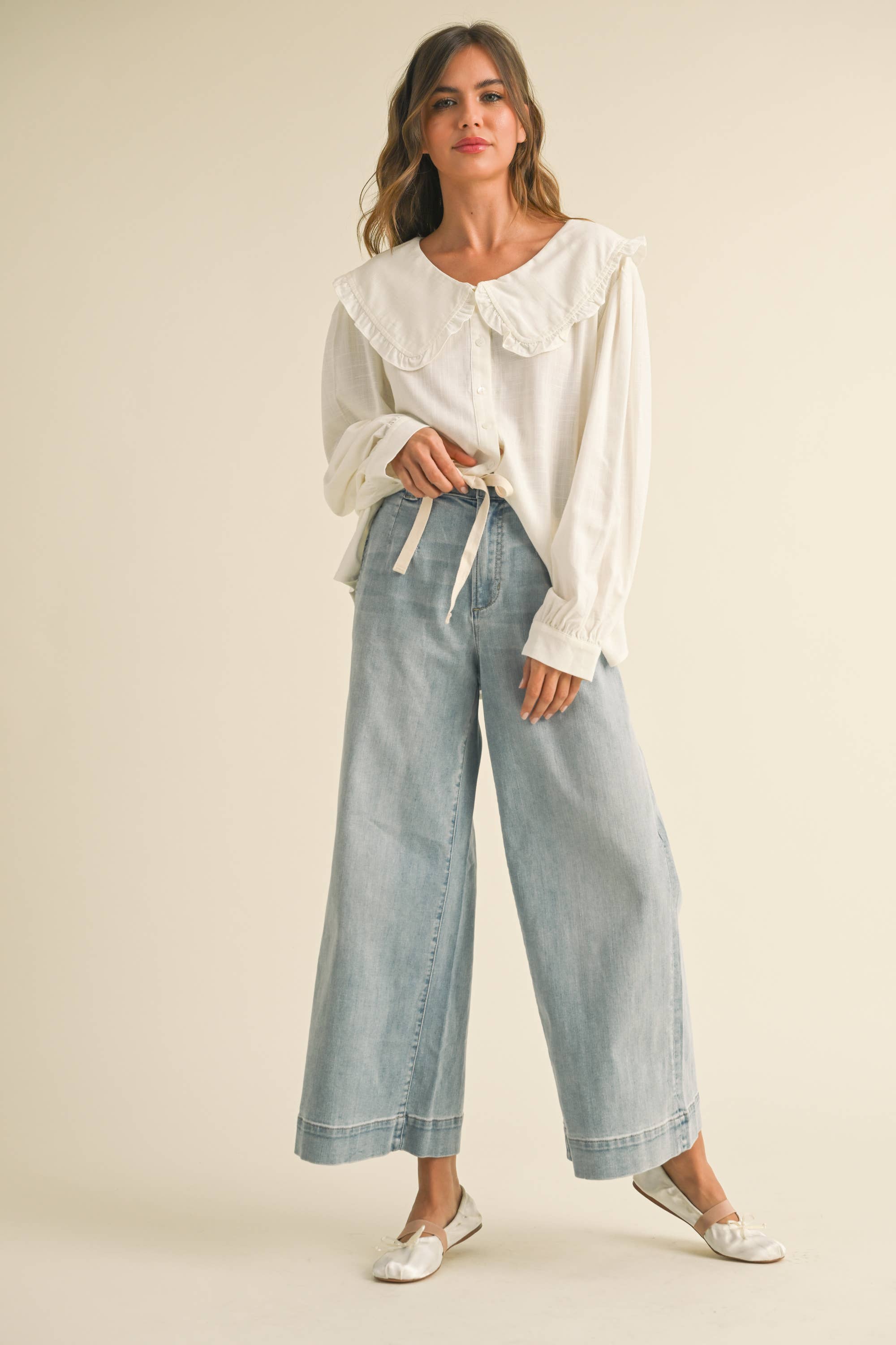 Woman wearing wide-leg light denim pants with a tie-front waist, paired with a white blouse and ballet flats.