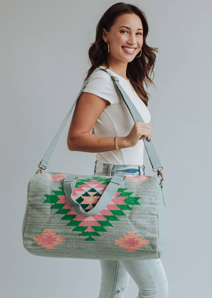 Sage green weekender bag with pink, emerald, and peach geometric tribal pattern, held by a woman in light-wash jeans