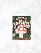 A greeting card with a hand-painted illustration of a mushroom and the text 'love you so mush' on a background that resembles a garden.