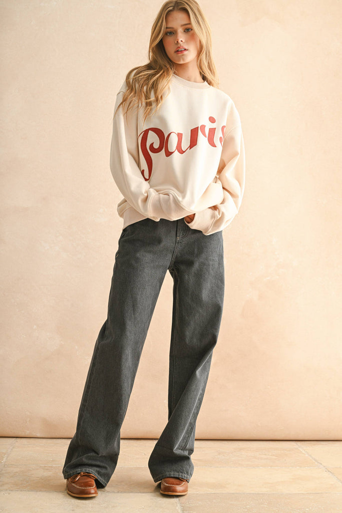 Woman wearing a cream sweater with bold red embroidered “Paris” lettering, paired with dark denim jeans.