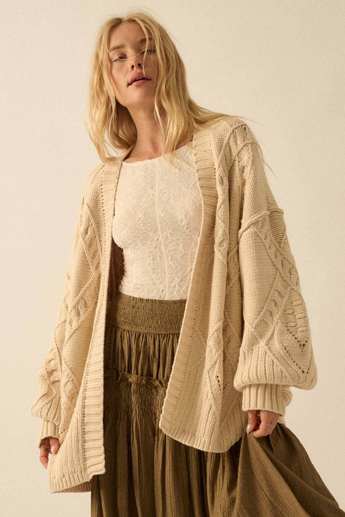 Woman wearing a beige cable knit cardigan over a white top and brown skirt on a plain background