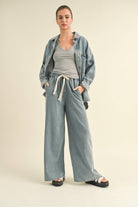 Model in an oversized denim jacket with front pockets. First image: jacket buttoned, paired with white shorts. Second image: jacket open, over a grey tank and denim pants.