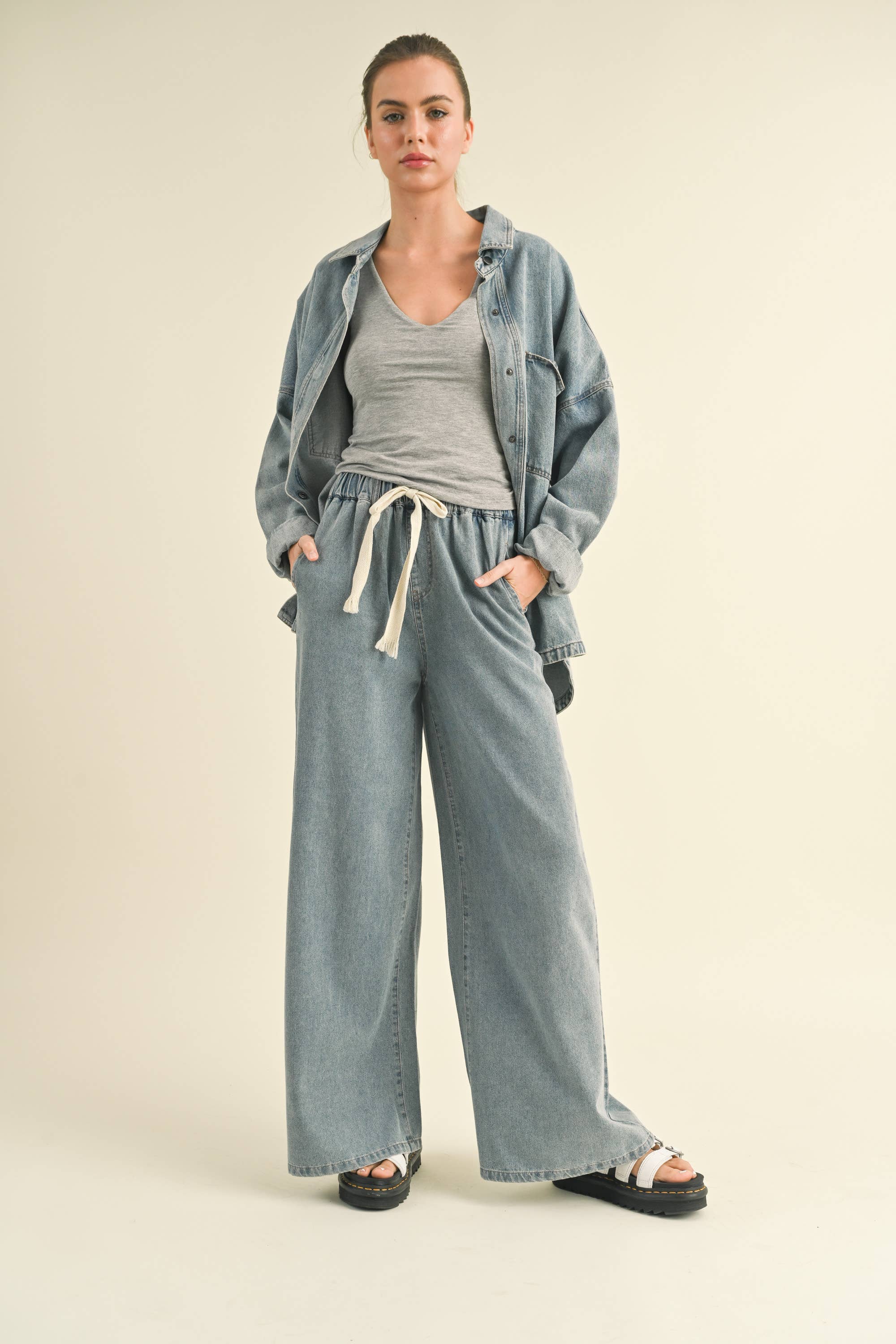 Model in an oversized denim jacket with front pockets. First image: jacket buttoned, paired with white shorts. Second image: jacket open, over a grey tank and denim pants.