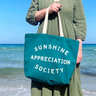 Teal tote bag with “Sunshine Appreciation Society” in white text, held by a person in a green dress at the beach