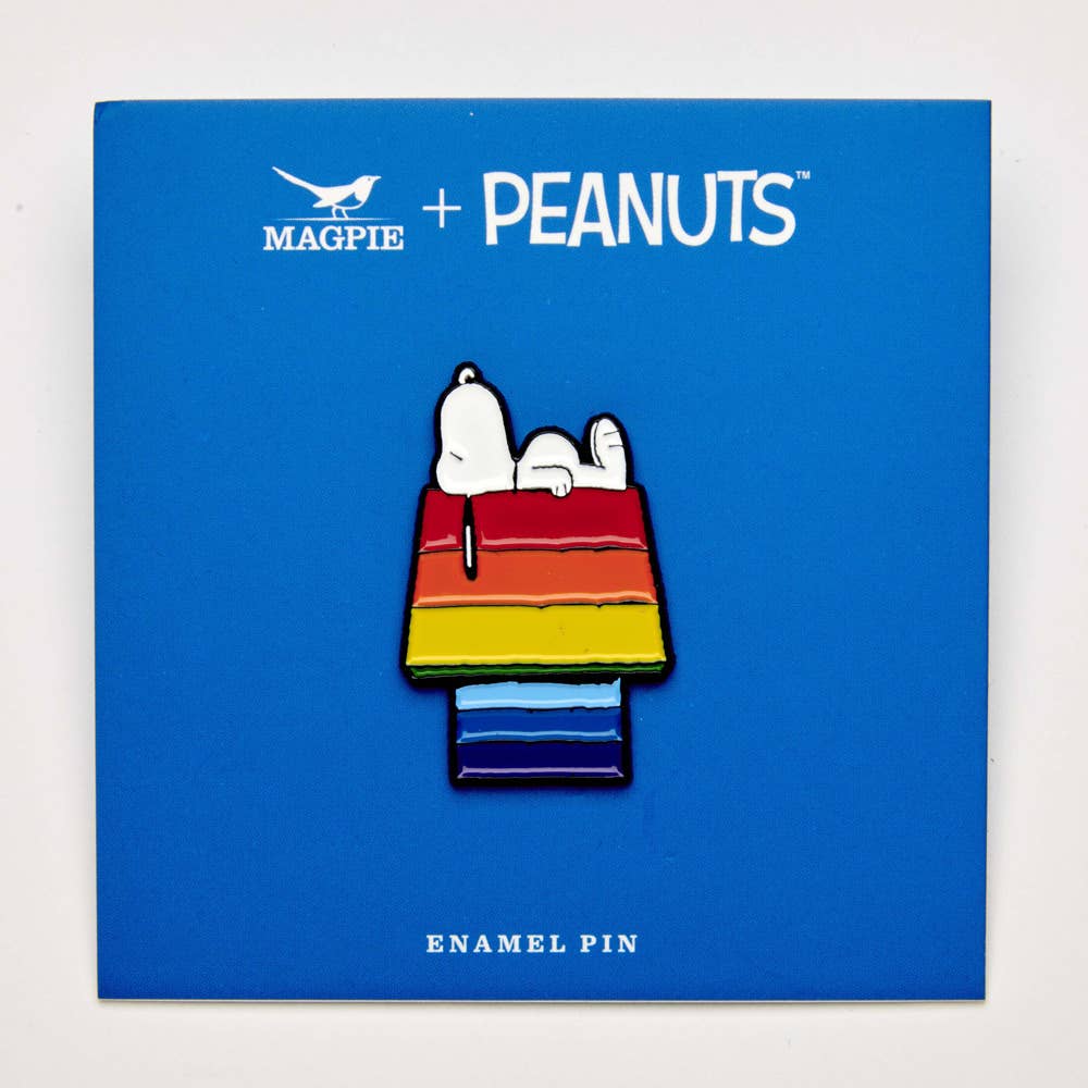 Enamel pin of Snoopy on a rainbow-striped doghouse displayed on blue packaging labeled "Magpie + Peanuts."