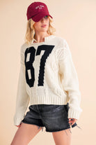 Woman wearing a cream knit sweater with a bold black "87" graphic and dark denim shorts.