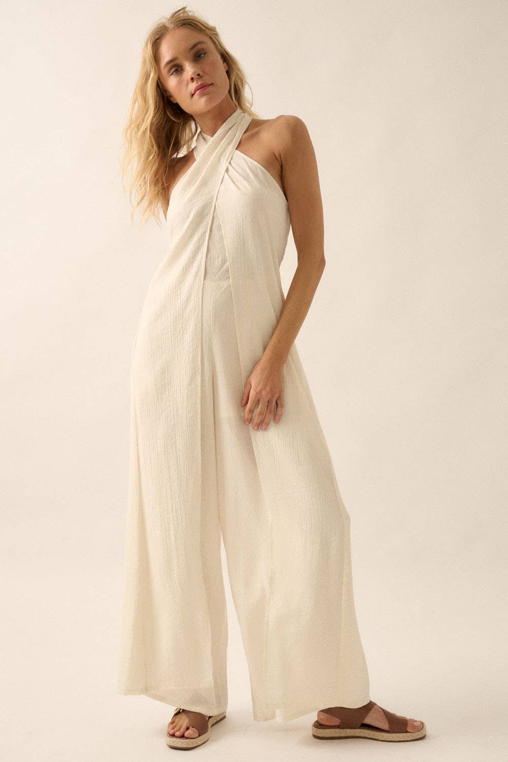 Woman wearing a white halter jumpsuit with tie-back neckline and wide-leg pants, styled with sandals.