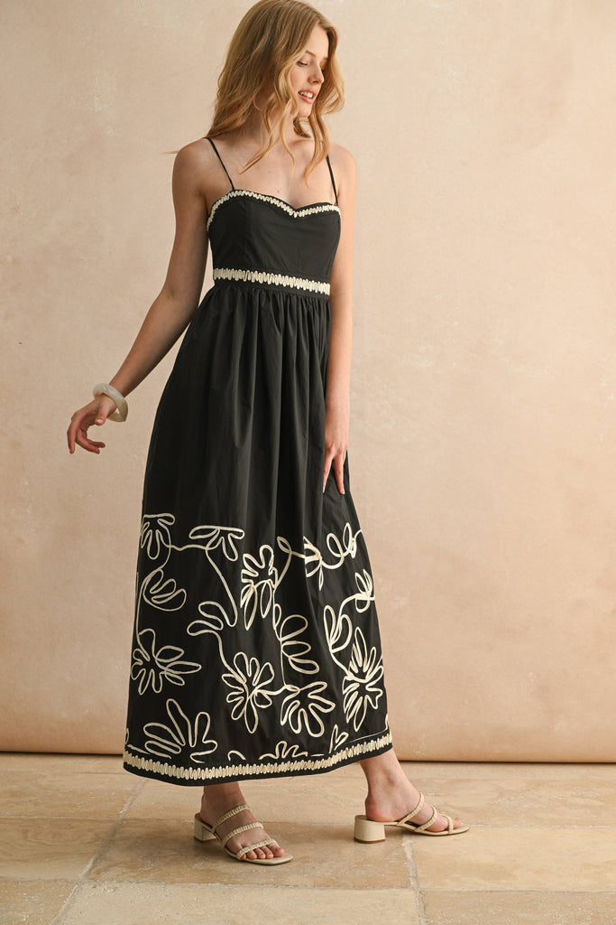 Woman wearing a black spaghetti-strap maxi dress with ivory floral embroidery and scalloped trim detail.