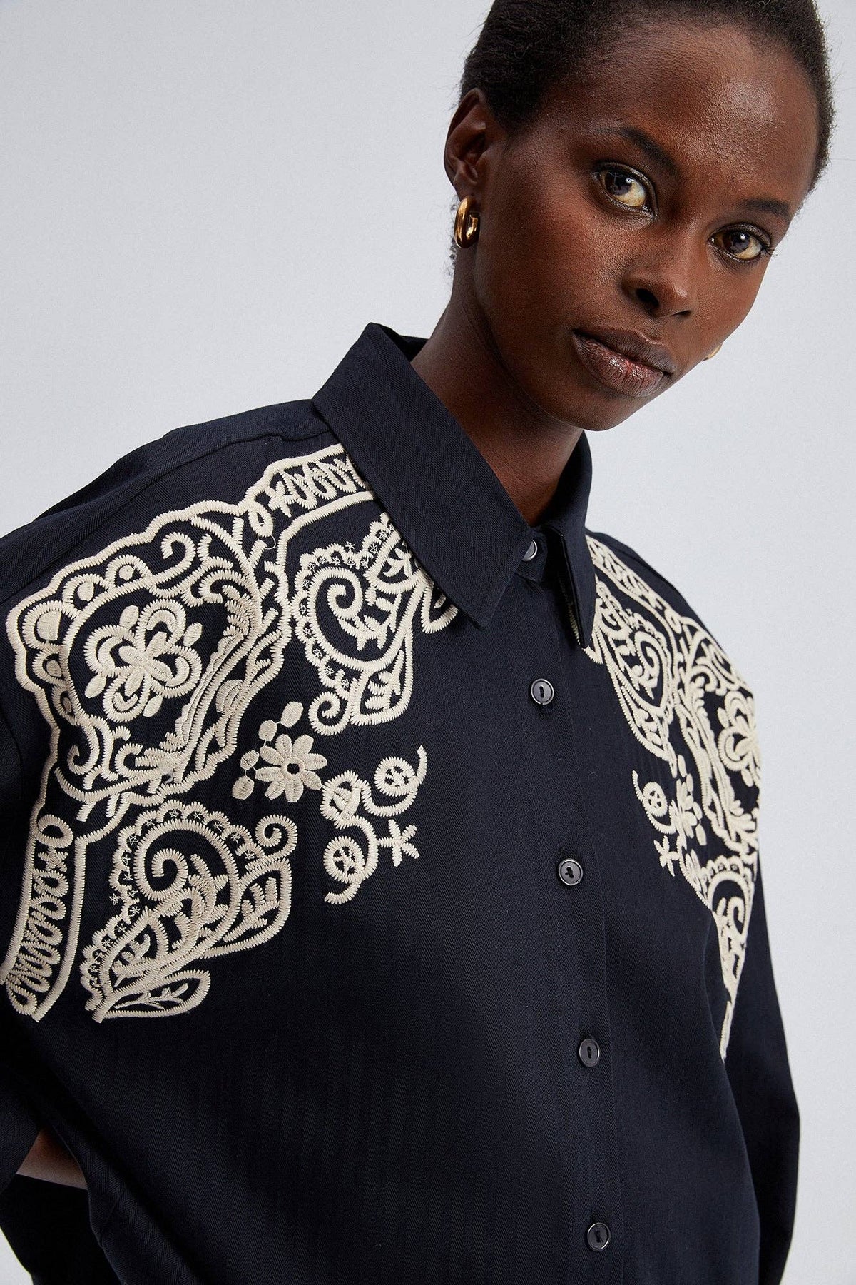 Model wearing black oversized shirt with cream embroidery on shoulders and front yoke.