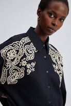 Model wearing black oversized shirt with cream embroidery on shoulders and front yoke.