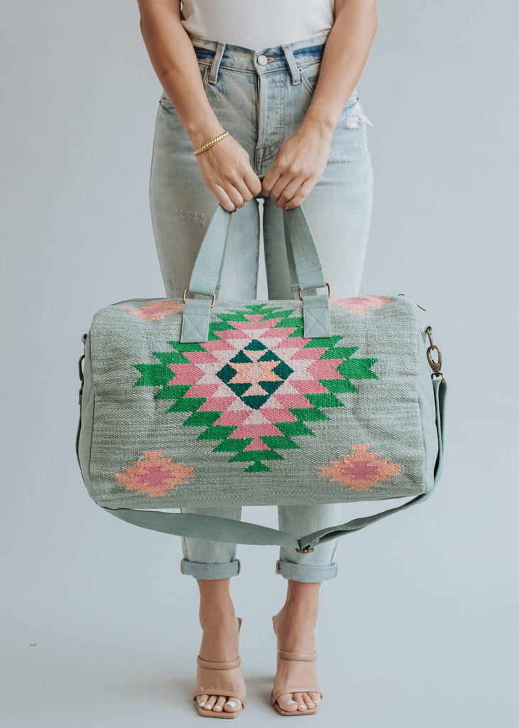 Sage green weekender bag with pink, emerald, and peach geometric tribal pattern, held by a woman in light-wash jeans