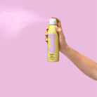 Hand holding Bask SPF 50 Sunscreen Spray, showing the spray function against a sunny beach scene.