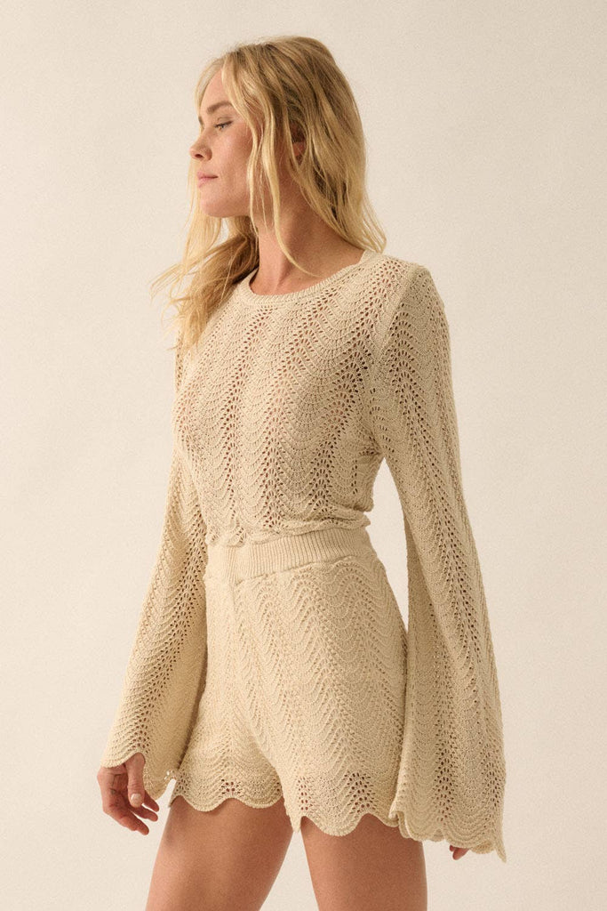 Woman wearing a beige knit romper with bell sleeves, scalloped hems, and open-weave detailing.