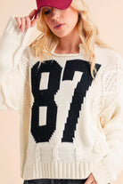 Woman wearing a cream knit sweater with a bold black "87" graphic and dark denim shorts.