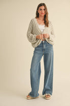 Woman wearing a loose cable knit cardigan with wide sleeves, paired with a white ribbed tank and blue denim jeans.