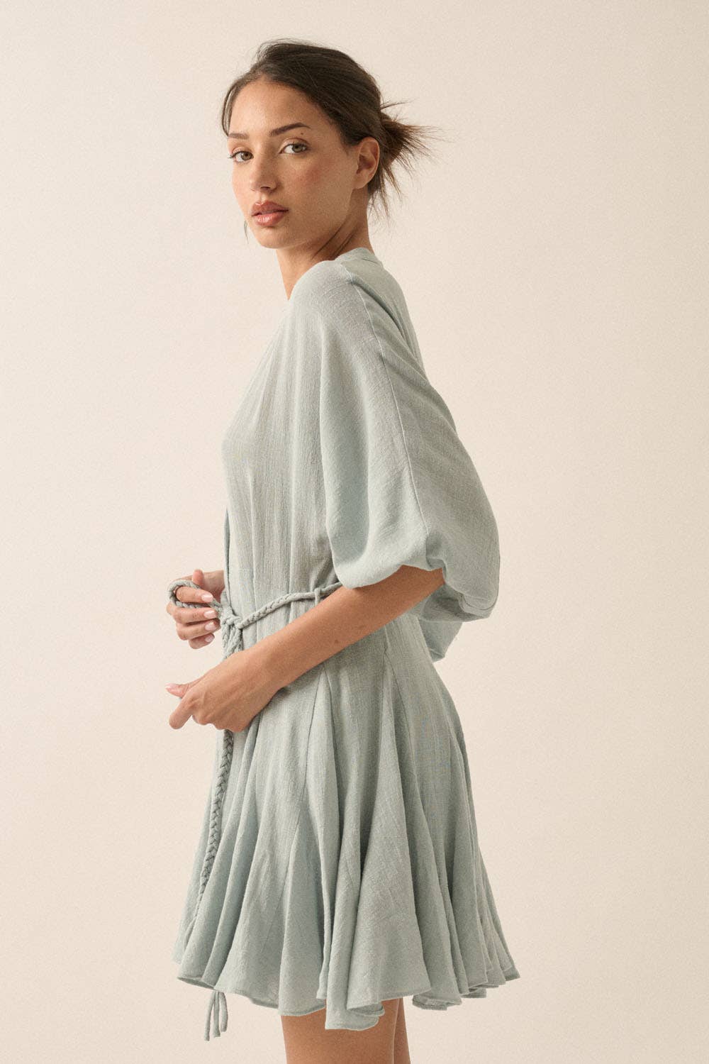 Woman wearing a sage green button-down mini dress with a braided waist tie and flowy hemline.