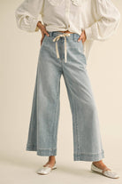 Woman wearing wide-leg light denim pants with a tie-front waist, paired with a white blouse and ballet flats.