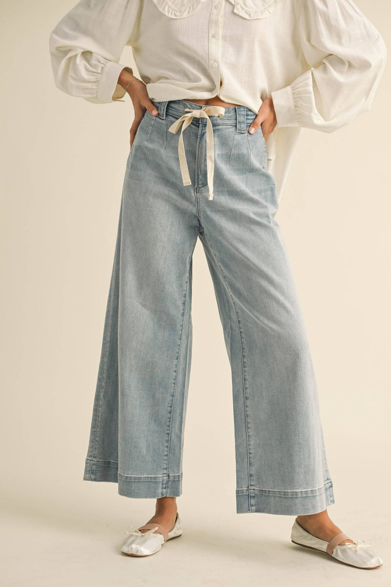Woman wearing wide-leg light denim pants with a tie-front waist, paired with a white blouse and ballet flats.
