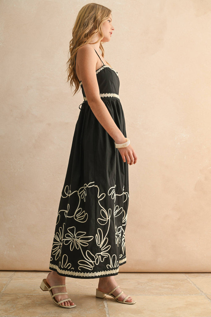 Woman wearing a black spaghetti-strap maxi dress with ivory floral embroidery and scalloped trim detail.