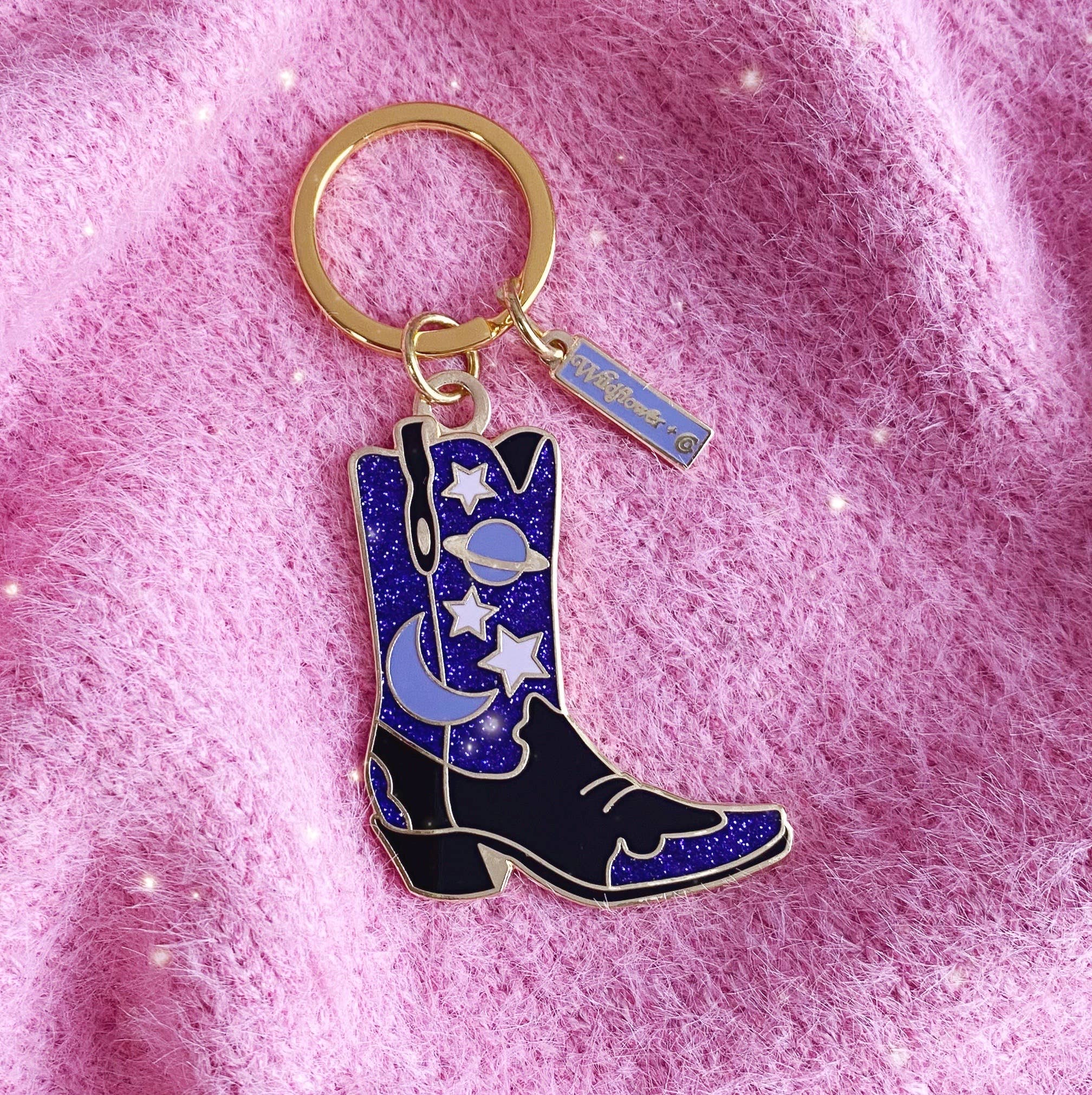 Celestial Cowgirl Boot Enamel Keychain: A stylish keychain featuring a black and blue cowgirl boot with celestial designs, including stars, moons, and planets, set against a pink background.