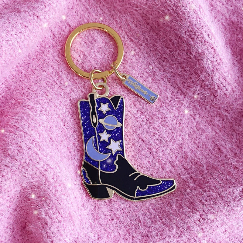 Celestial Cowgirl Boot Enamel Keychain: A stylish keychain featuring a black and blue cowgirl boot with celestial designs, including stars, moons, and planets, set against a pink background.