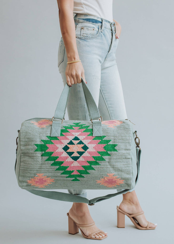 Sage green weekender bag with pink, emerald, and peach geometric tribal pattern, held by a woman in light-wash jeans