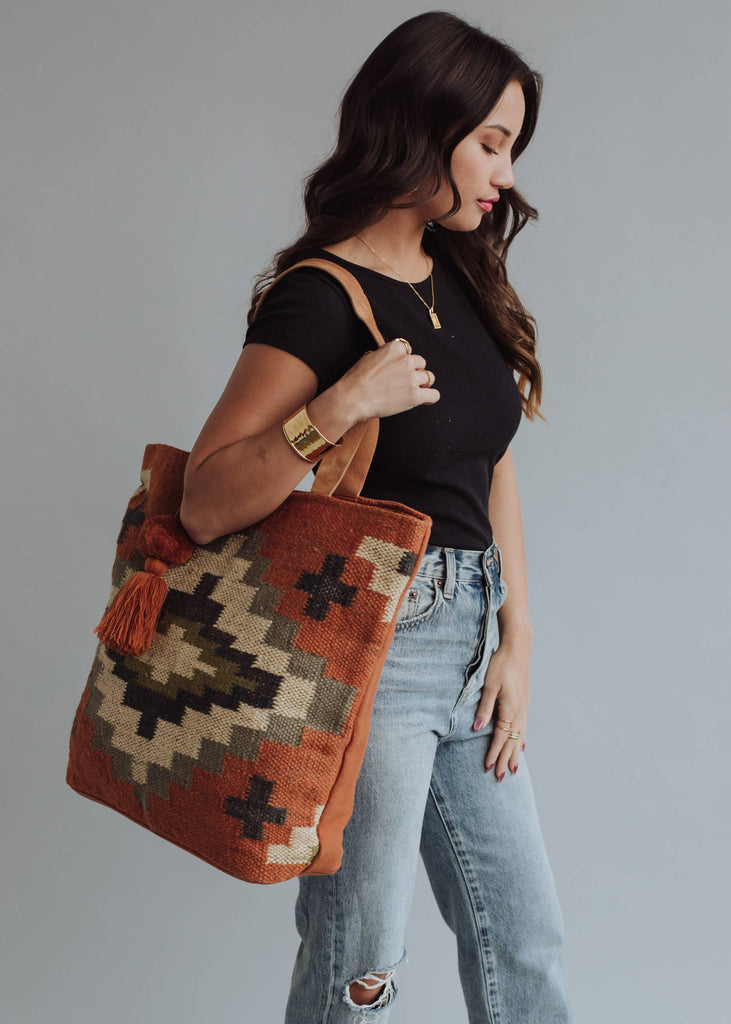 Woman holding a rust and olive green diamond-patterned woven tote with tassel