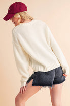 Woman wearing a cream knit sweater with a bold black "87" graphic and dark denim shorts.