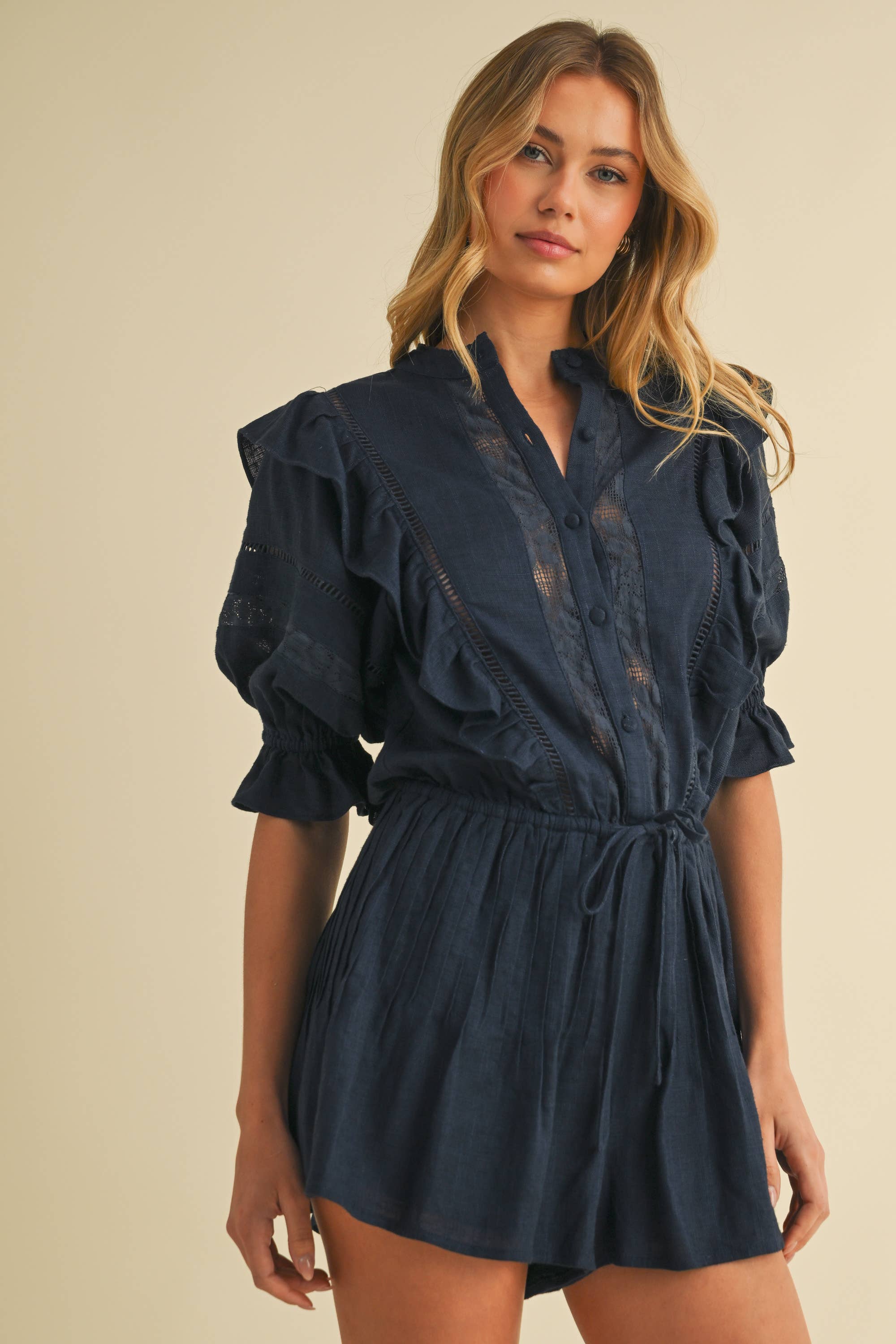 Woman wearing a navy cotton romper with puff sleeves, ruffle and lace details, and a drawstring waist.