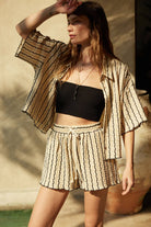 Woman wearing a beige striped two-piece lounge set with button-up shirt and matching drawstring shorts.