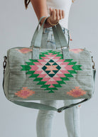 Sage green weekender bag with pink, emerald, and peach geometric tribal pattern, held by a woman in light-wash jeans