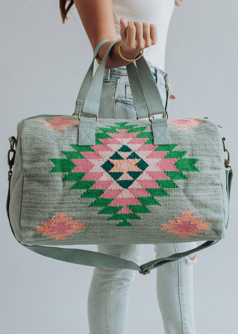 Sage green weekender bag with pink, emerald, and peach geometric tribal pattern, held by a woman in light-wash jeans